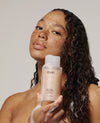 Woman with curly wet hair holding OUAI Thick Hair Shampoo, 10 fl oz bottle.