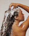 Woman with curly hair lathering shampoo under shower water, side profile view.