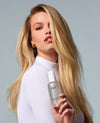 A blonde woman in a white ribbed mock-neck top holds a silver and white OUAI hair product bottle against a solid light gray background.