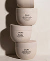 Three OUAI hair treatment jars stacked vertically on a white surface against a white wall, with dramatic side shadows. The top jar is partially cropped at the top edge.