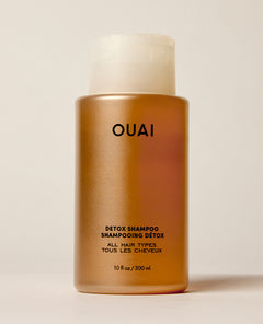 An amber OUAI shampoo bottle stands upright on a warm cream surface beneath a translucent cap. Text: “OUAI”, “DETOX SHAMPOO”, “SHAMPOOING DÉTOX”, “ALL HAIR TYPES”, “TOUS LES CHEVEUX”, “10 fl oz / 300 ml”.