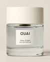 A short clear perfume bottle containing pale blue liquid stands on a cream studio surface beneath a wide white cap. Text: “OUAI”, “DEAN STREET”, “EAU DE PARFUM”.