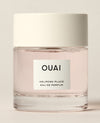 A short clear perfume bottle containing pale pink liquid stands on a warm cream studio surface beneath a wide white cap. Text: “OUAI”, “MELROSE PLACE”, “EAU DE PARFUM”.