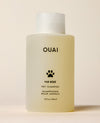 A translucent OUAI Fur Bébé Pet Shampoo bottle containing pale yellow liquid stands against a cream studio background. Text: “OUAI”, “FUR BÉBÉ”, “PET SHAMPOO”, “