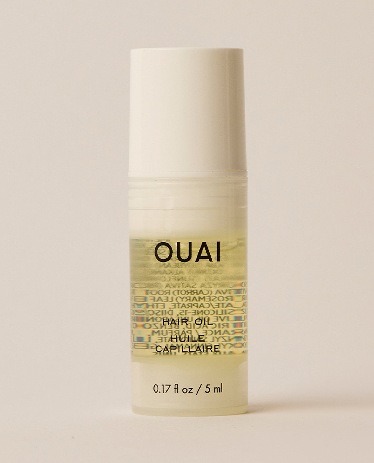 A small cylindrical OUAI Hair Oil bottle containing pale golden liquid stands upright on a cream surface, topped with a matte white cap.