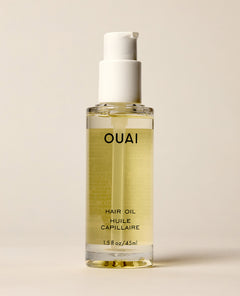A clear OUAI Hair Oil pump bottle containing pale yellow liquid stands against a softly lit cream background. Text: “OUAI”, “HAIR OIL”, “HUILE CAPILLAIRE”, “1.5 fl oz / 45 ml”.