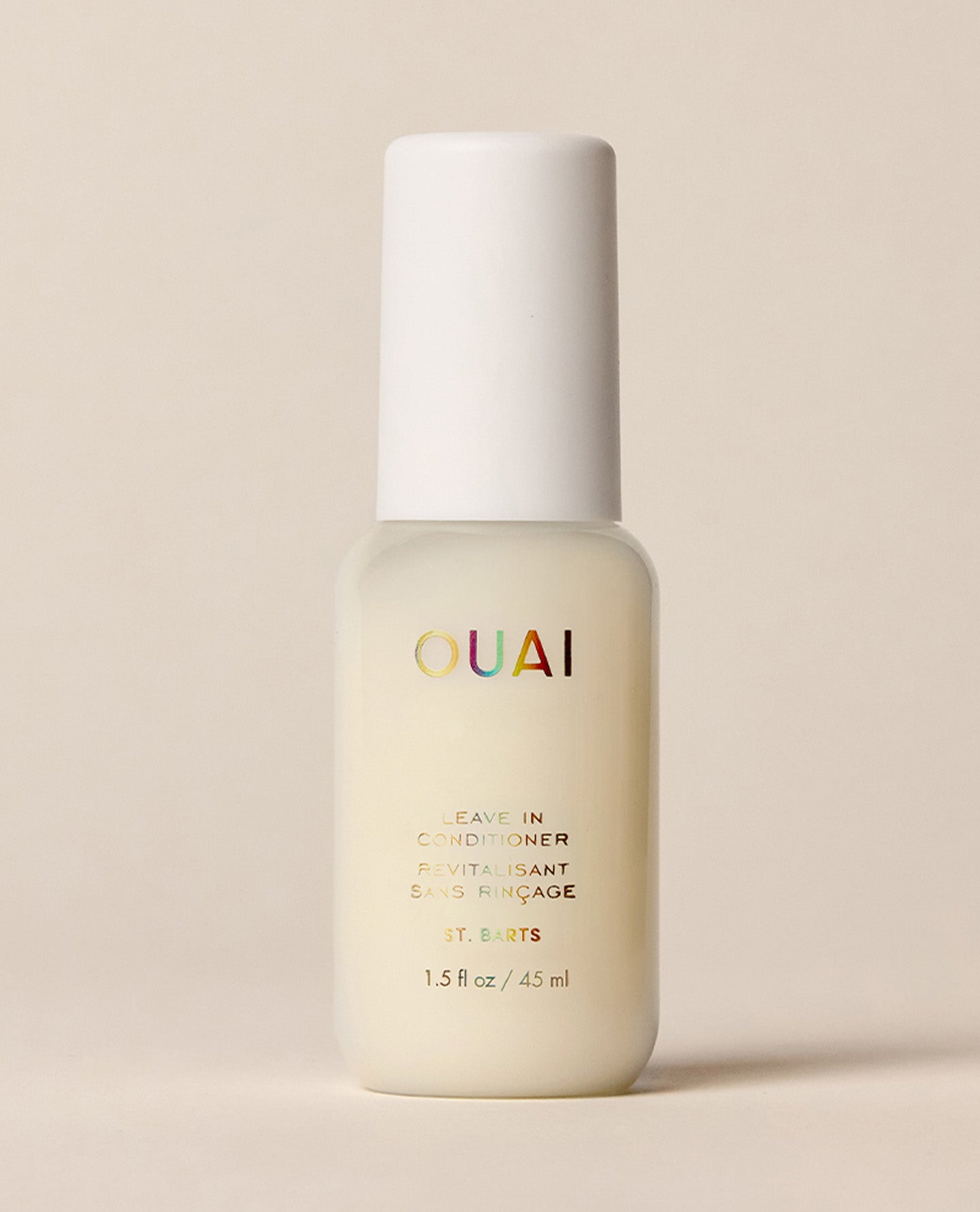 A larger frosted OUAI St. Barts Leave-In Conditioner travel bottle stands upright against a softly lit cream background, finished with iridescent branding and a rounded white cap.