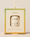 A boxed OUAI St. Barts solid fragrance ornament hangs by a cream cord, with the compact visible through a square window against a warm neutral backdrop.