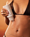 A close-up of a person's midsection wearing a black bikini, holding a white tube of OUAI skin care and squeezing a clear gel onto the skin against a warm orange-brown background.