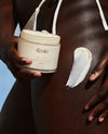 A close-up of a person holding a jar of OUAI body cream, with a smear of the white cream on their skin