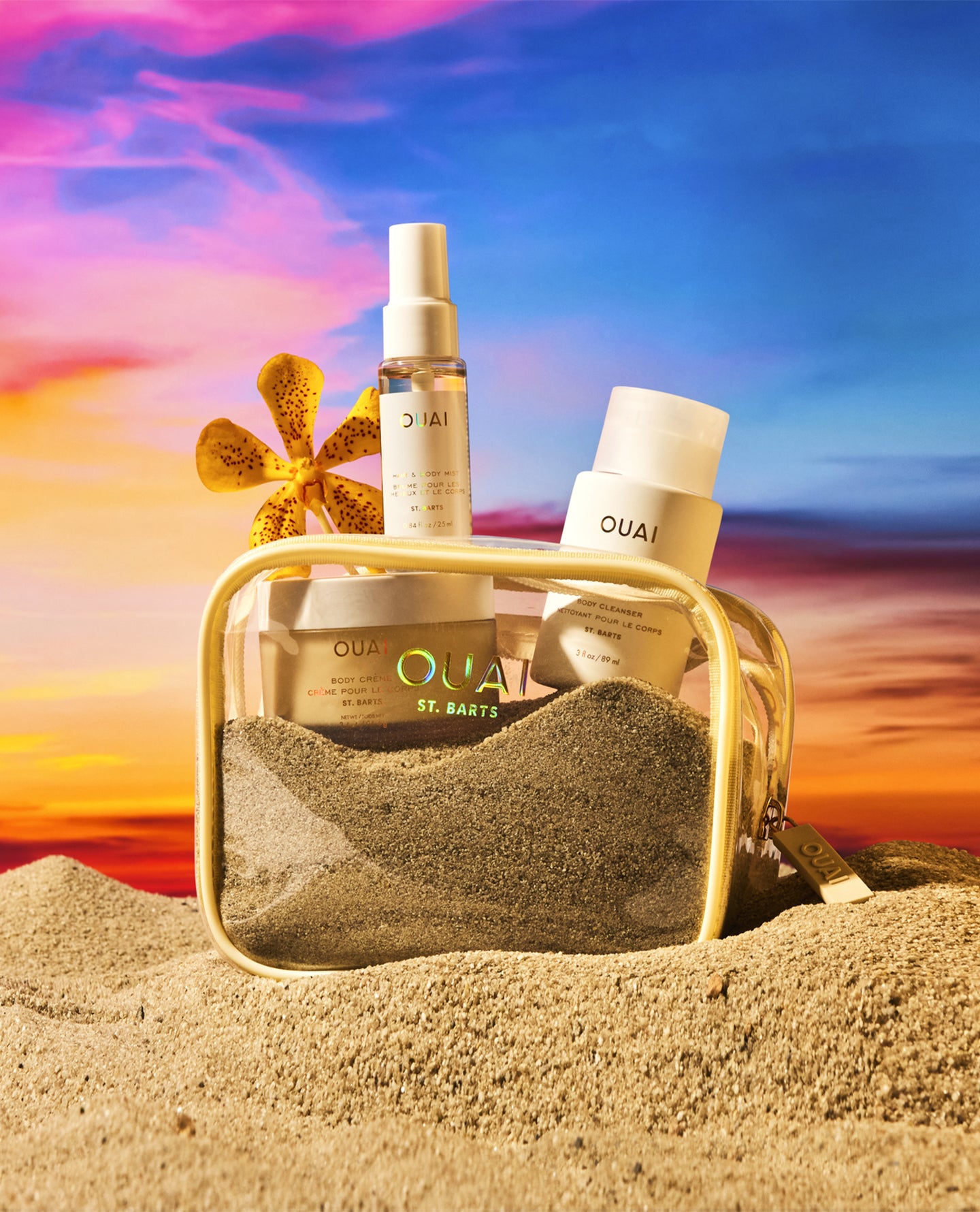 OUAI St. Bart's beach-themed skincare set arranged in a clear zippered pouch filled with sand, with a gold frame, set against a vibrant beach sunset sky and sand dunes.